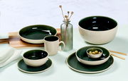 Vielseitiges Geschirrset in elegantem Dunkelgrün und Creme, bestehend aus Tassen, Tellern und Schalen, auf einem Tisch arrangiert. Seitliche Perspektive.