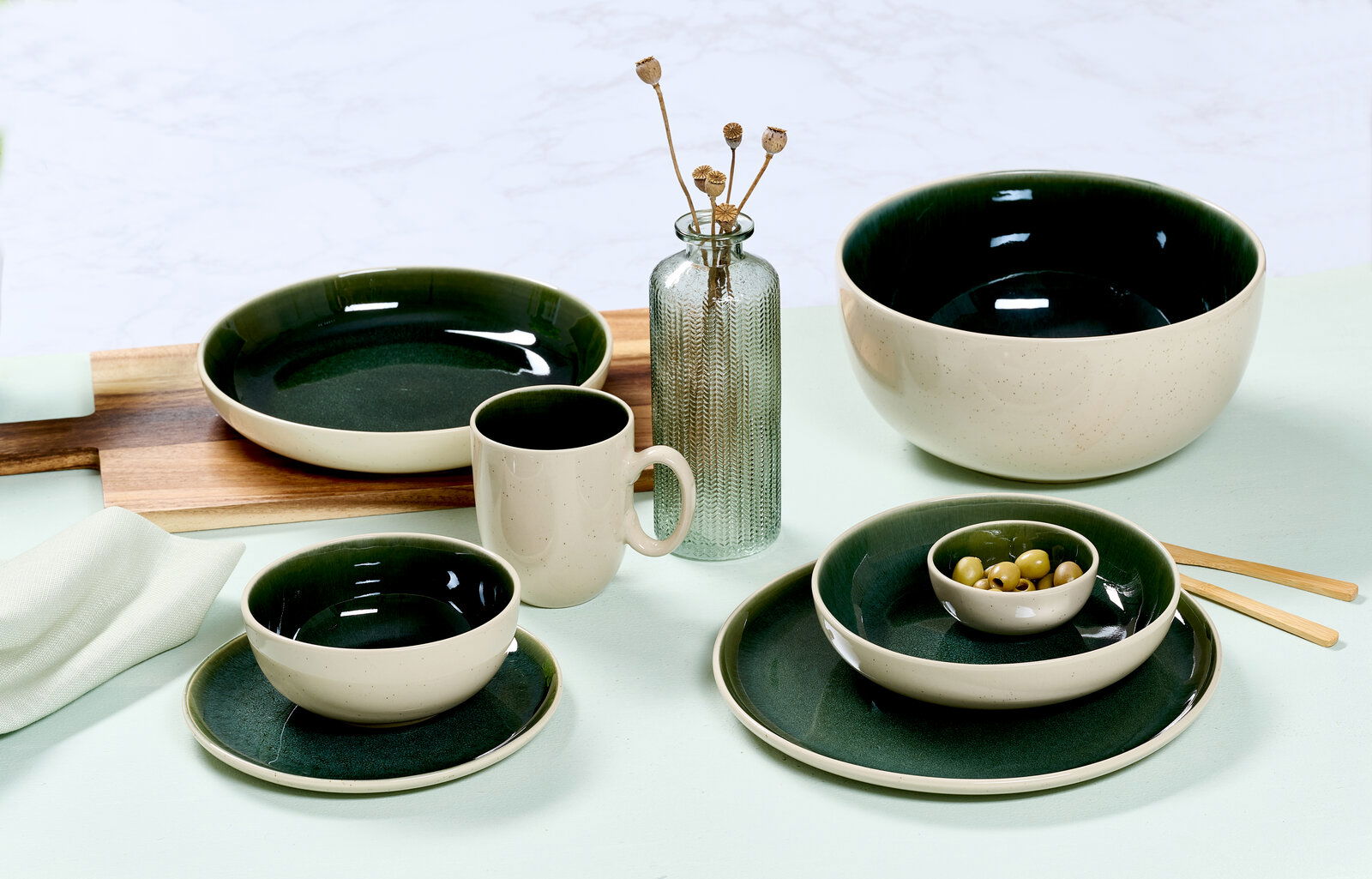 Vielseitiges Geschirrset in elegantem Dunkelgrün und Creme, bestehend aus Tassen, Tellern und Schalen, auf einem Tisch arrangiert. Seitliche Perspektive.