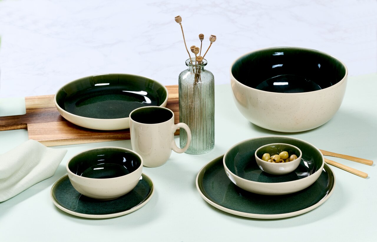 Vielseitiges Geschirrset in elegantem Dunkelgrün und Creme, bestehend aus Tassen, Tellern und Schalen, auf einem Tisch arrangiert. Seitliche Perspektive.