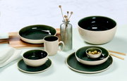 Vielseitiges Geschirrset in elegantem Dunkelgrün und Creme, bestehend aus Tassen, Tellern und Schalen, auf einem Tisch arrangiert. Seitliche Perspektive.
