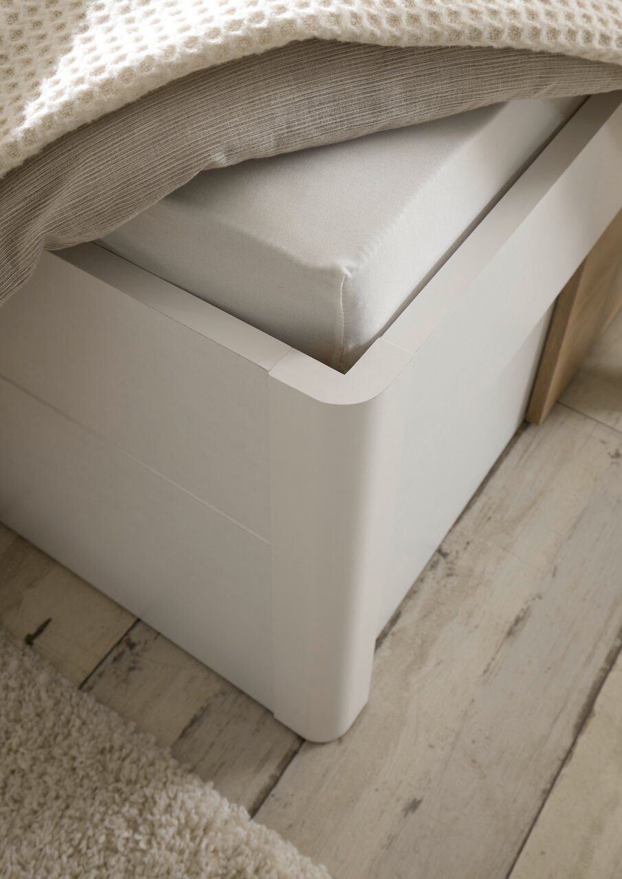 Nahaufnahme einer weißen Bettrahmenecke mit Matratze und Decke, aus der Perspektive von oben.