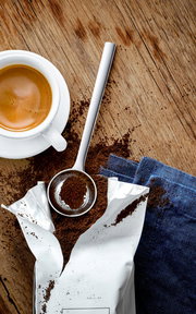Kaffeemaßlöffel aus Edelstahl, gefüllt mit gemahlenem Kaffee, neben einer Tasse Espresso auf einem Holztisch, von oben betrachtet.
