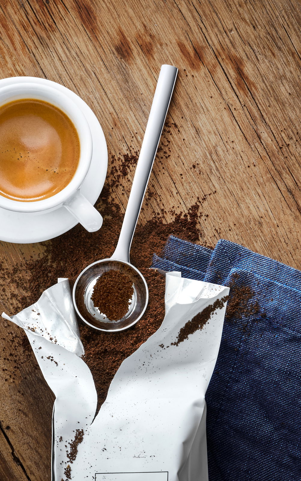 Kaffeemaßlöffel aus Edelstahl, gefüllt mit gemahlenem Kaffee, neben einer Tasse Espresso auf einem Holztisch, von oben betrachtet.