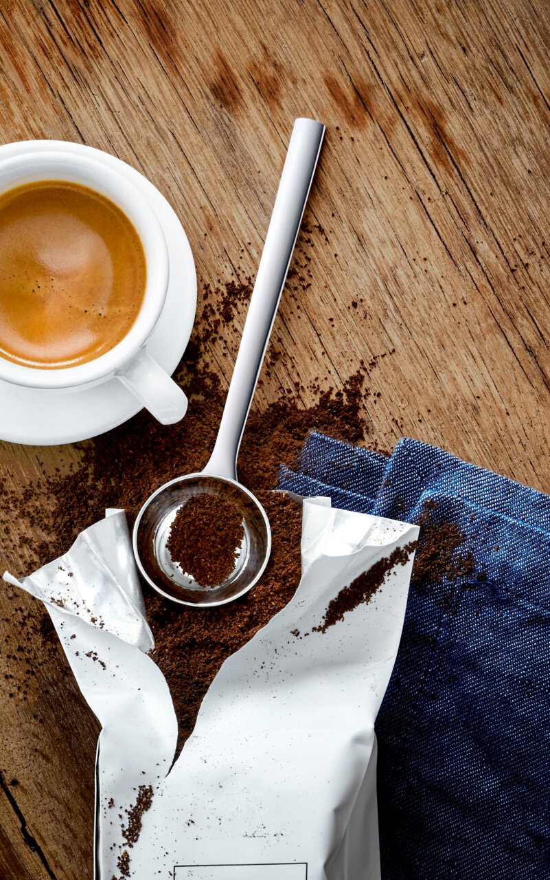 Een roestvrijstalen koffielepel gevuld met gemalen koffie, naast een kopje espresso op een houten tafel, gezien van bovenaf.