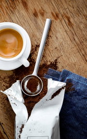 Kaffeemaßlöffel aus Edelstahl, gefüllt mit gemahlenem Kaffee, neben einer Tasse Espresso auf einem Holztisch, von oben betrachtet.