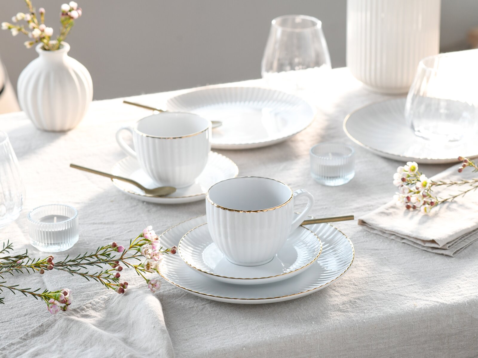 Schlichtes weißes Porzellan-Kaffeegeschirr mit geriffelter Struktur und feinem Goldrand, bestehend aus Tassen mit Untertassen und Tellern, dekorativ auf einem hellen Leinentischtuch gedeckt; Perspektive: leicht erhöhte, schräge Seitenansicht von vorn.