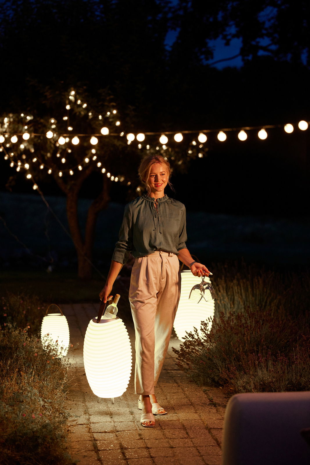 Frau trägt eine leuchtende Akku-Leuchte und Lautsprecher in einem Garten bei Nacht, umgeben von Lichterketten, aus der Perspektive von vorne.