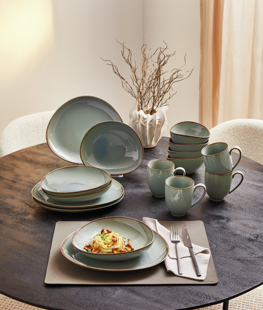 Een 16-delig servies in zachtgroen op een ronde tafel, bestaande uit borden, schalen en kopjes, met een decoratief takje op de achtergrond. Aanzicht van bovenaf.