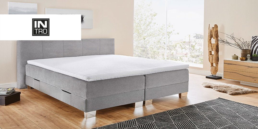 Een modern grijs gestoffeerd bed met een wit matras op een houten vloer met een grijs-wit tapijt.