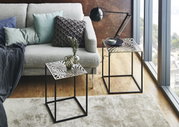 Wohnzimmer-Setting mit zwei modernen Beistelltischen aus schwarzem Metall mit dekorativer, blattförmig ausgeschnittener Tischplatte; links ein Tisch mit Glasvasen vor einem grauen Sofa, rechts ein Tisch neben bodentiefem Fenster mit Lampe und Schale – Per