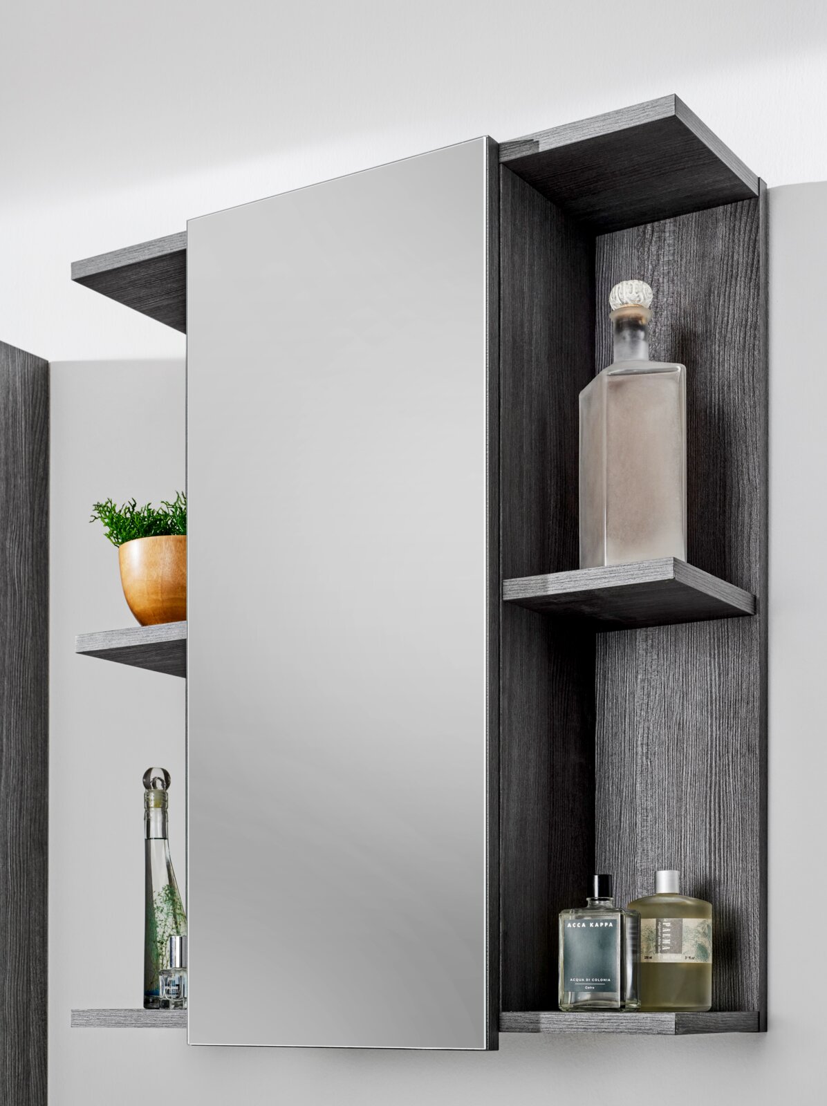 Badezimmerspiegelschrank mit grauer Holzoptik, seitlichen offenen Regalen und großem Spiegel, bestückt mit Flaschen und Deko; Frontansicht mit leicht seitlicher Perspektive.