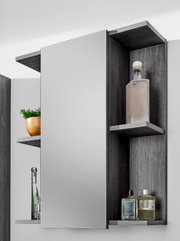 Badezimmerspiegelschrank mit grauer Holzoptik, seitlichen offenen Regalen und großem Spiegel, bestückt mit Flaschen und Deko; Frontansicht mit leicht seitlicher Perspektive.