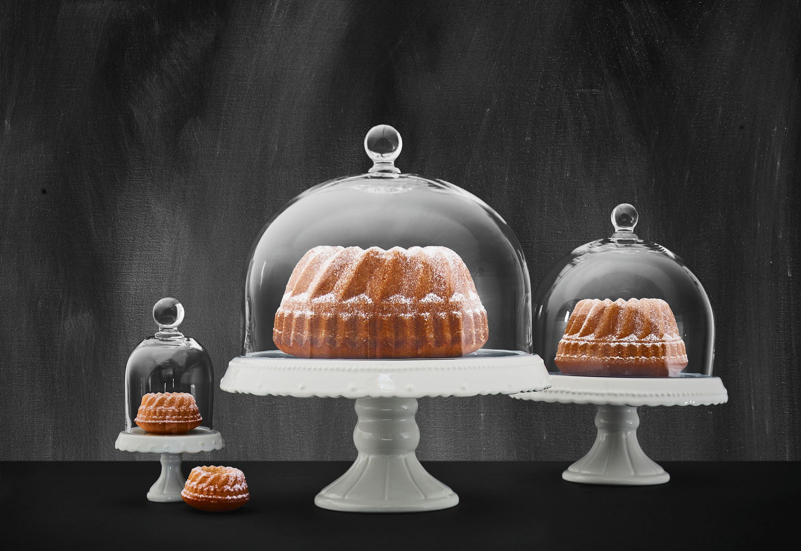 Birkmann Glashaube VINTAGE Drei unterschiedlich große Glashauben auf weißen Tortenständern, die von vorne gezeigt werden, mit Kuchen darunter.