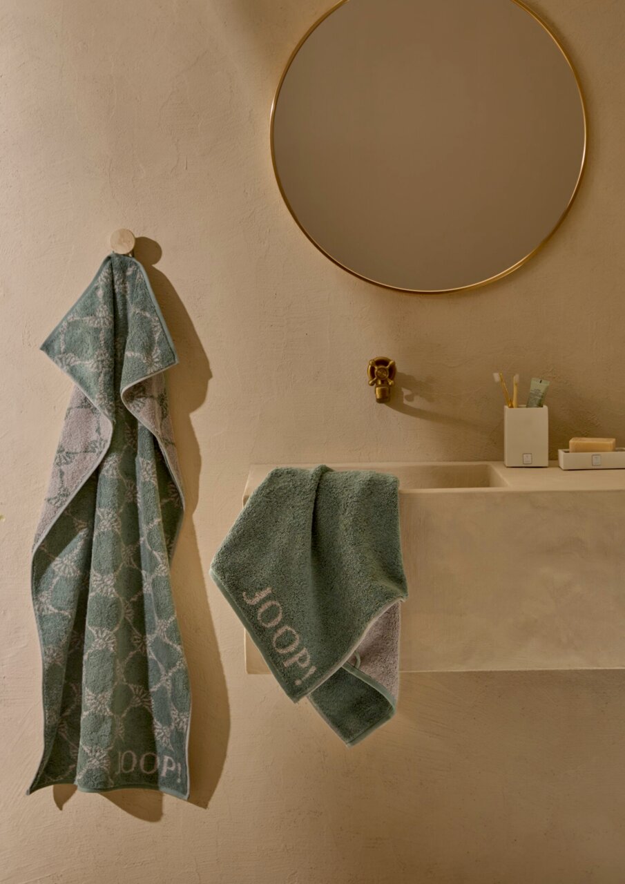 Zwei grüne Handtücher mit Muster, eines hängt an einem Haken, das andere liegt auf einem Waschbecken. Perspektive: Badezimmeransicht mit Spiegel und Waschbecken.