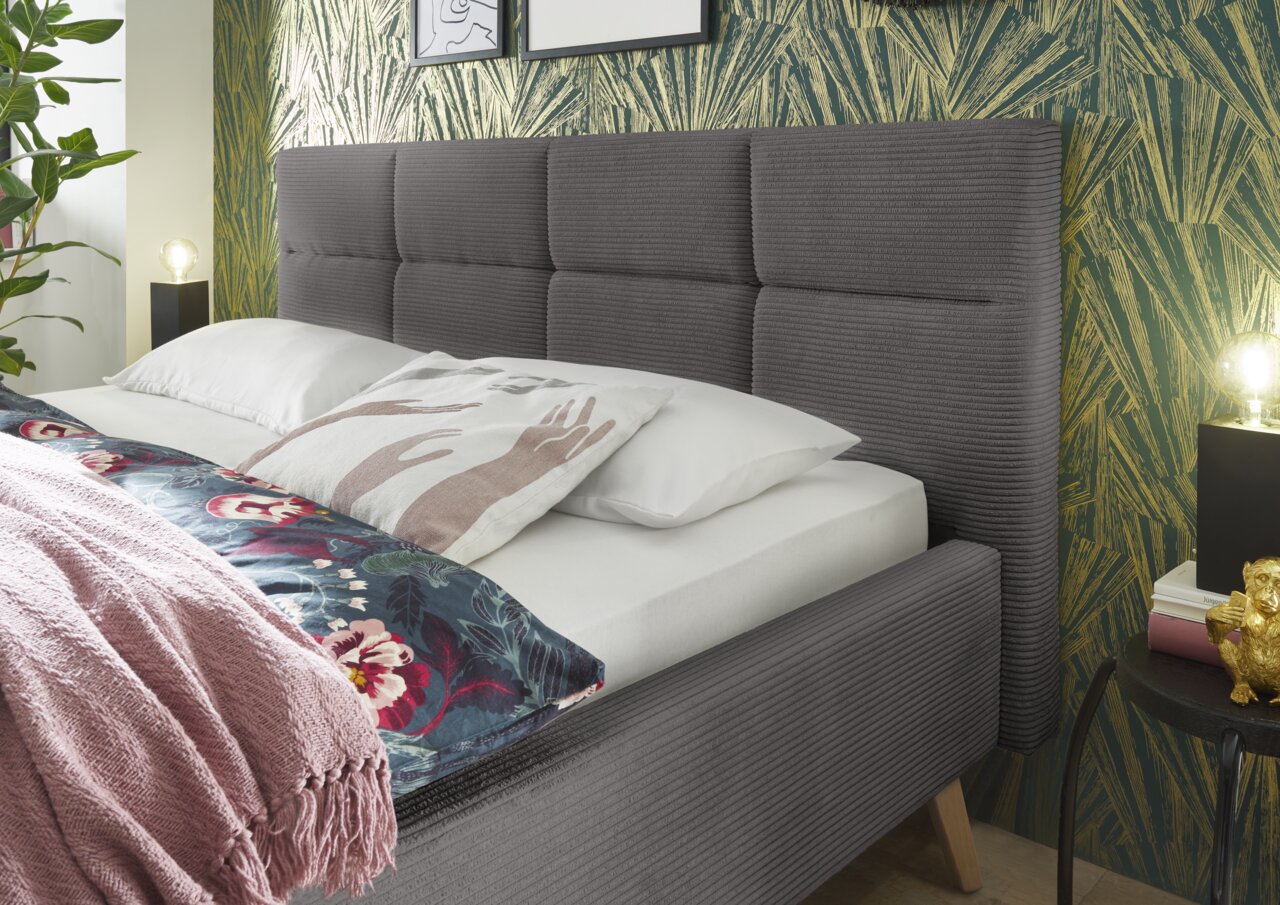 Graues Polsterbett mit gestepptem Kopfteil, seitliche Perspektive, dekoriert mit Kissen und Decke vor einer gemusterten Tapete.