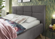 Graues Polsterbett mit gestepptem Kopfteil, seitliche Perspektive, dekoriert mit Kissen und Decke vor einer gemusterten Tapete.