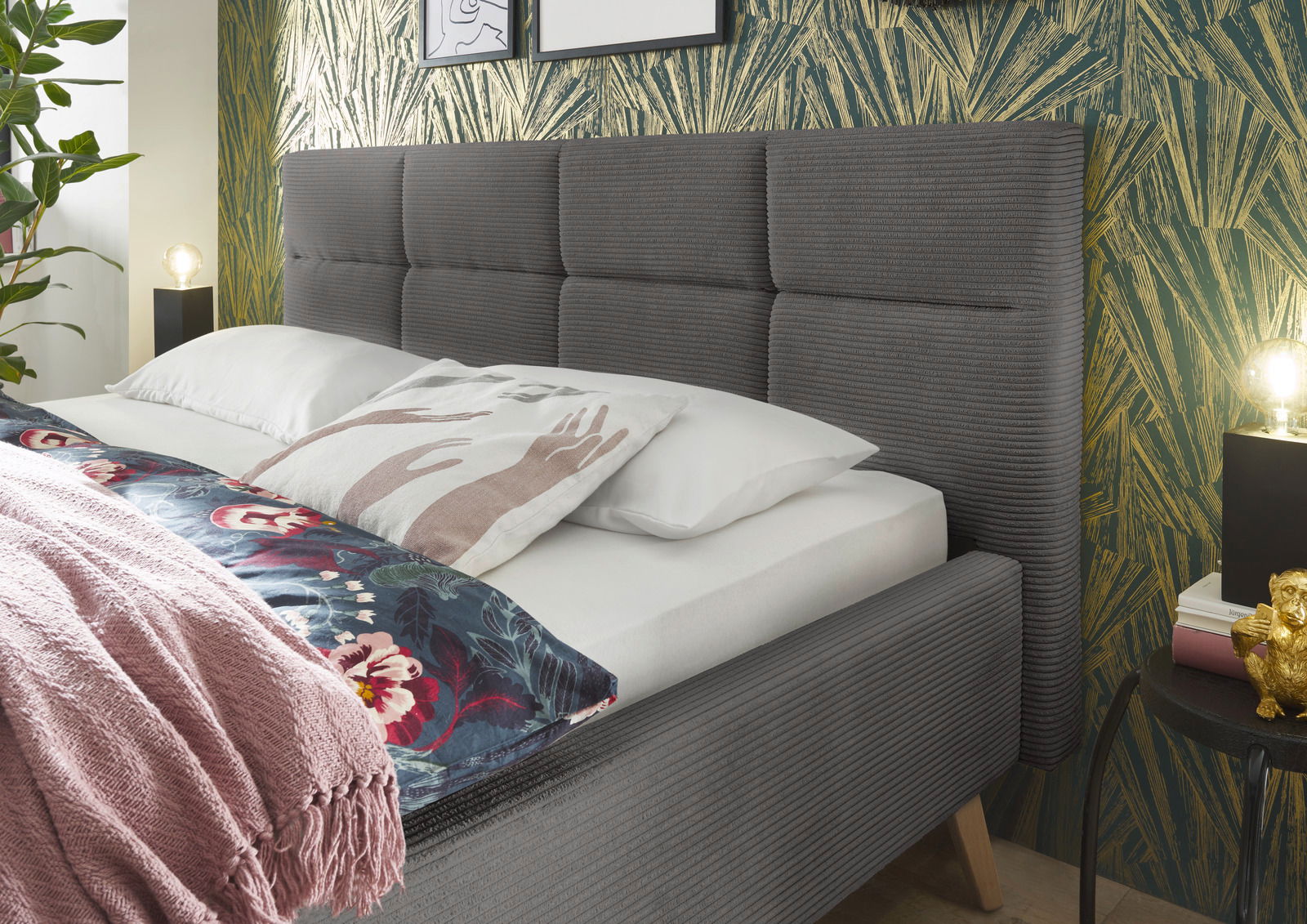 Graues Polsterbett mit gestepptem Kopfteil, seitliche Perspektive, dekoriert mit Kissen und Decke vor einer gemusterten Tapete.