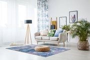 Moderner Wohnraum mit einem blauen Teppich in der Mitte, umgeben von einem beigen Sofa, Pflanzen und dekorativen Kissen, aus der Perspektive eines stehenden Betrachters.