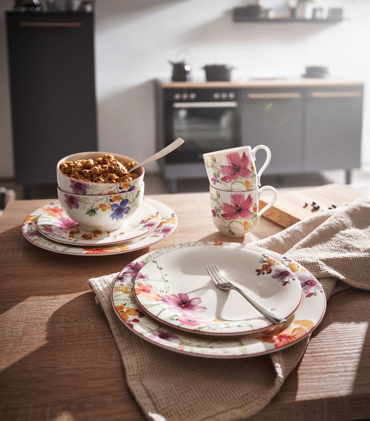 Frontalansicht eines eleganten Geschirrsets mit floralen Mustern, bestehend aus Tellern, Tassen und Schalen, auf einem Holztisch in einer modernen Küche.