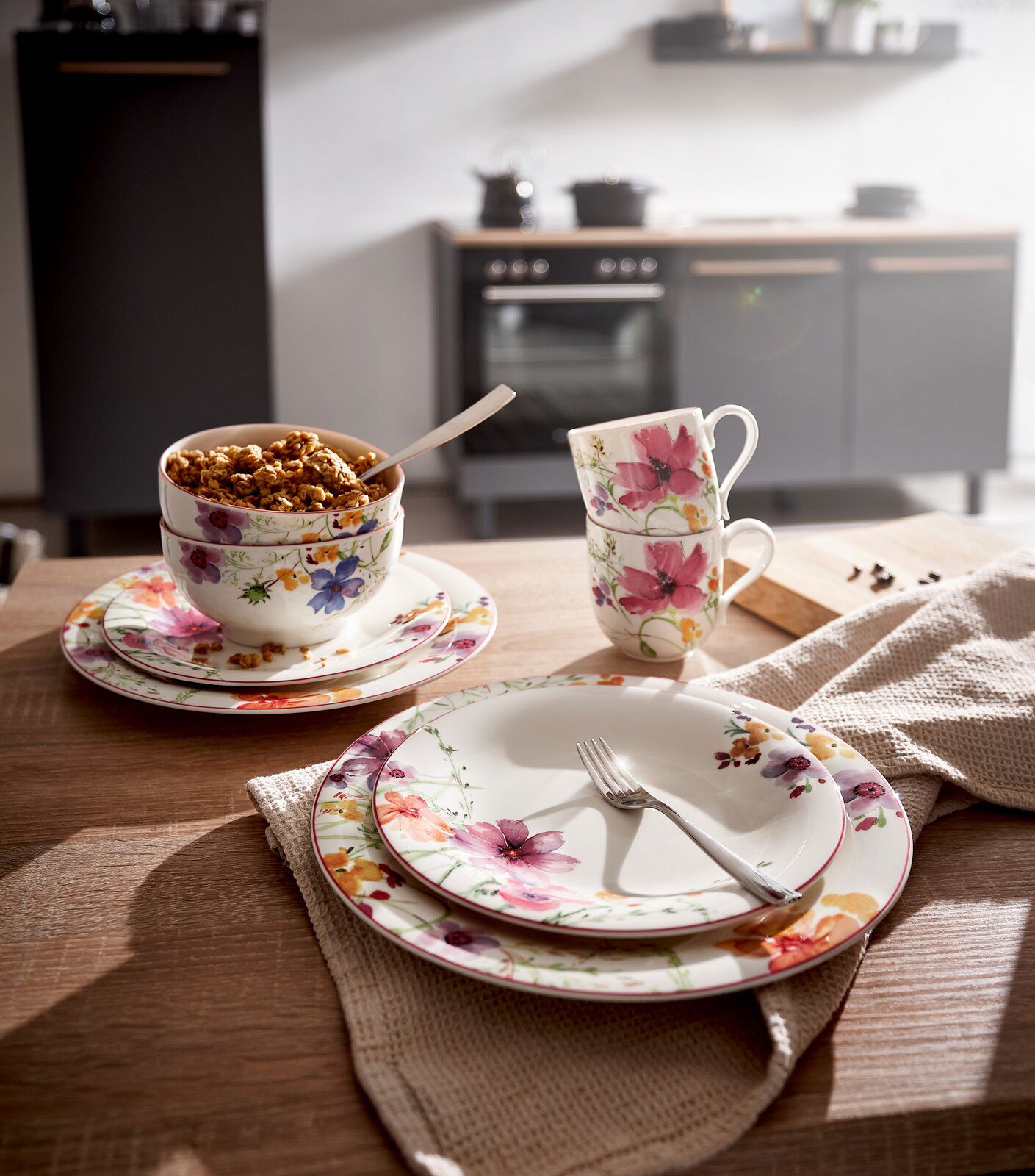 Frontalansicht eines eleganten Geschirrsets mit floralen Mustern, bestehend aus Tellern, Tassen und Schalen, auf einem Holztisch in einer modernen Küche.