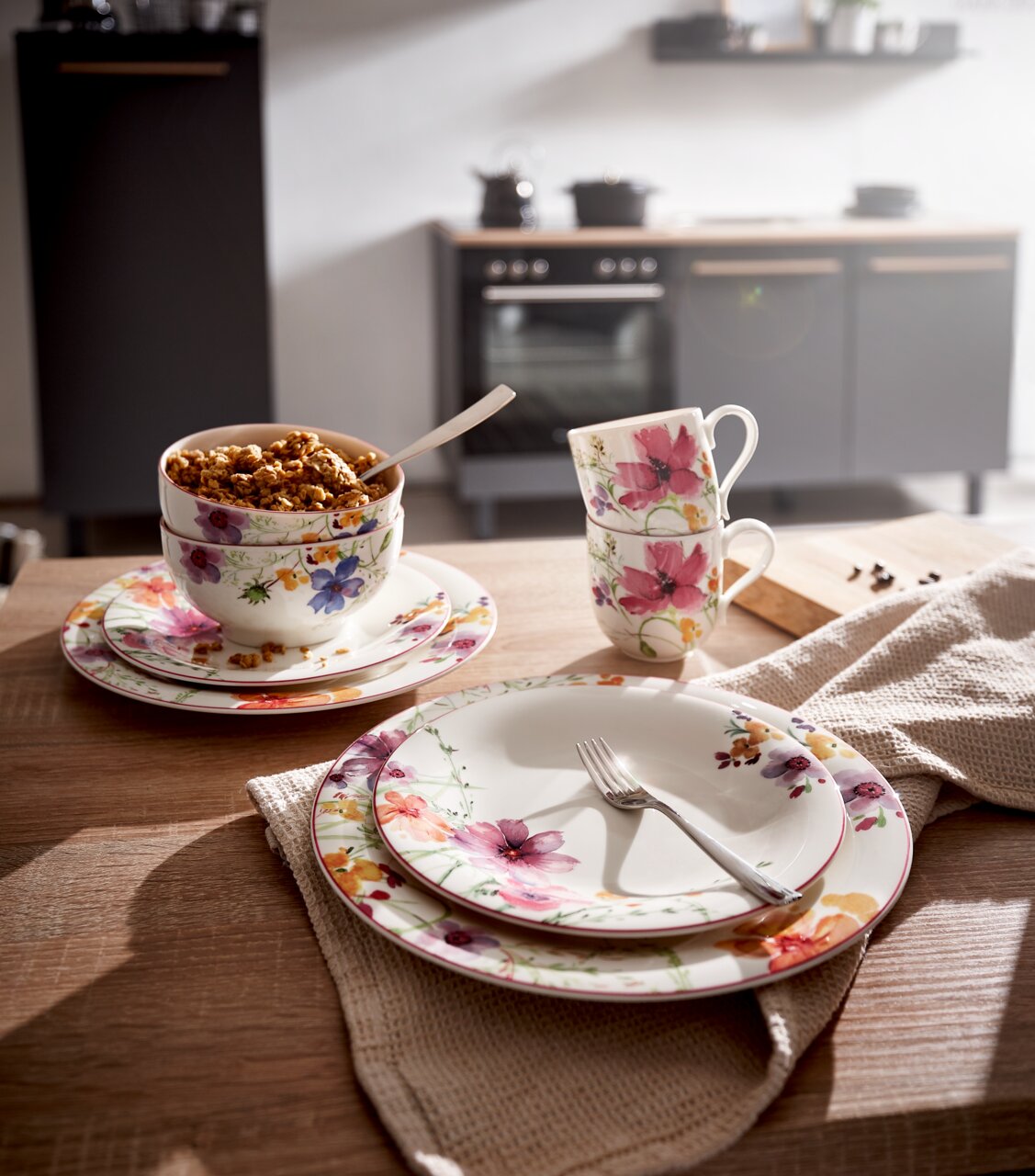 Frontalansicht eines eleganten Geschirrsets mit floralen Mustern, bestehend aus Tellern, Tassen und Schalen, auf einem Holztisch in einer modernen Küche.