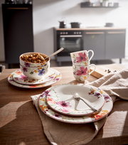 Frontalansicht eines eleganten Geschirrsets mit floralen Mustern, bestehend aus Tellern, Tassen und Schalen, auf einem Holztisch in einer modernen Küche.