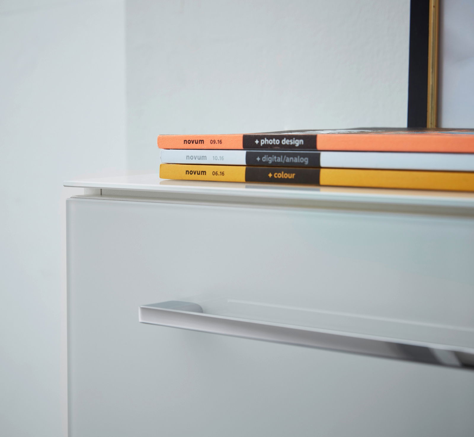 Nahaufnahme eines weißen Aktenschranks mit modernen Griffen, darauf liegen bunte Zeitschriften. Perspektive von schräg oben.