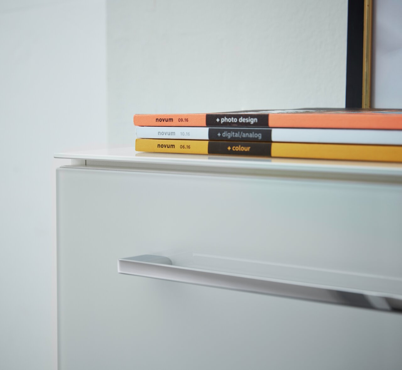 Close-up van een witte archiefkast met moderne handgrepen, met daarop kleurrijke tijdschriften. Schuin bovenaanzicht.