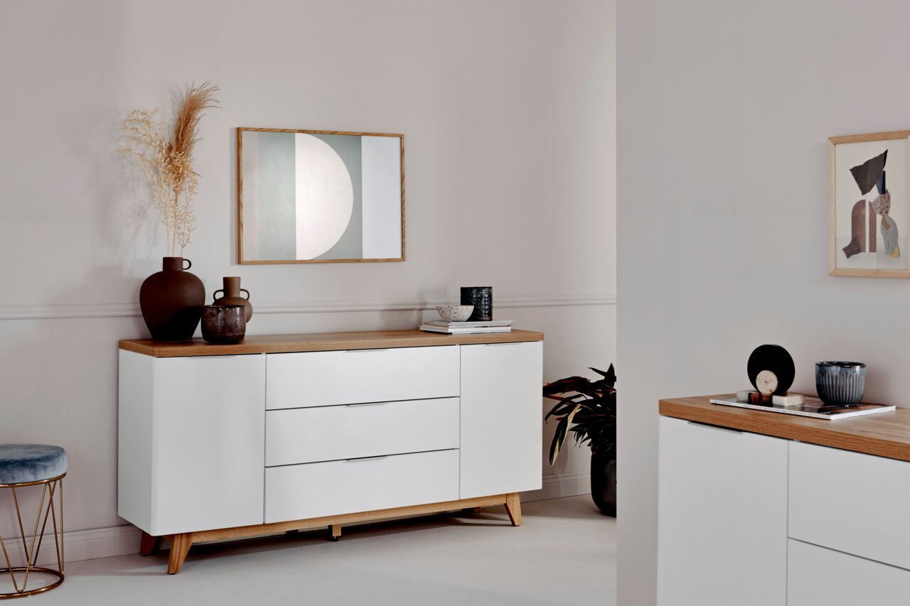 Weißes Sideboard mit Holzoberfläche, dekoriert mit Vasen und Kunstwerken, aus seitlicher Perspektive fotografiert.
