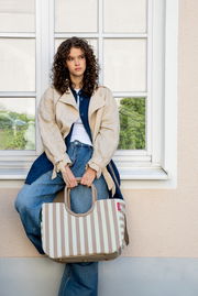 Beige-weiß gestreifte Tragetasche mit kurzen Henkeln, von vorn in leichter Seitenansicht fotografiert, gehalten von einer sitzenden Frau vor einem Fenster.
