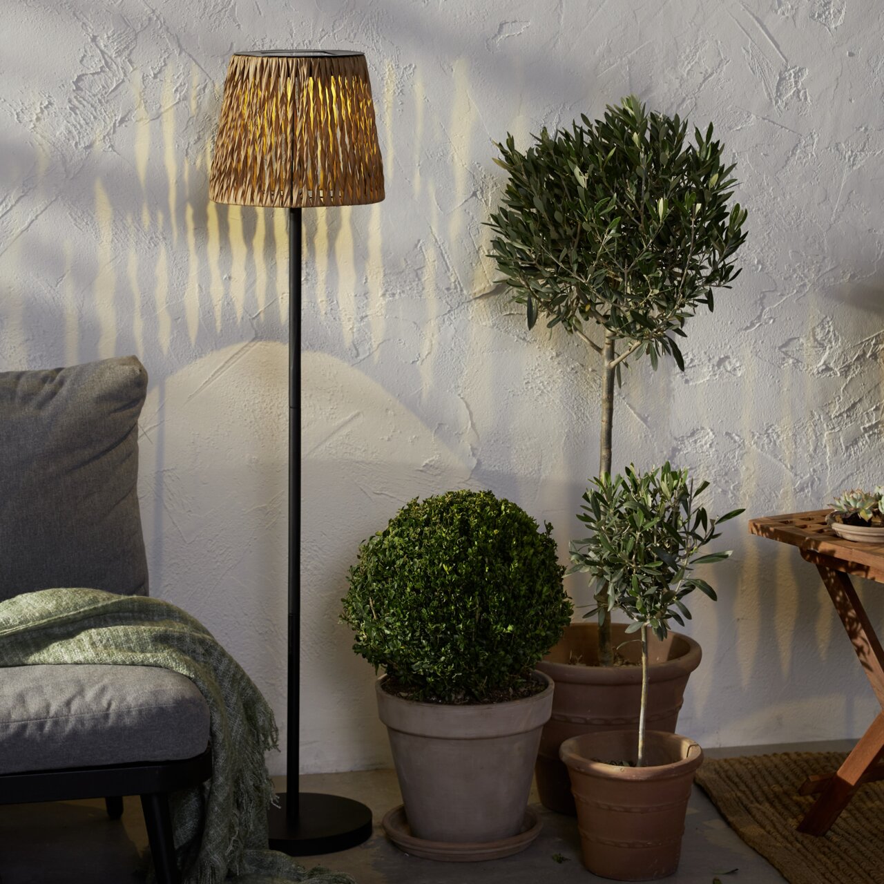 STAR TRADING LED-Solar-Stehleuchte CESTA Stehlampe mit geflochtenem Rattan-Lampenschirm und schwarzem Metallfuß neben Topfpflanzen vor strukturierter Wand, Innenraum-Szene, Perspektive: frontale Ansicht auf Augenhöhe.