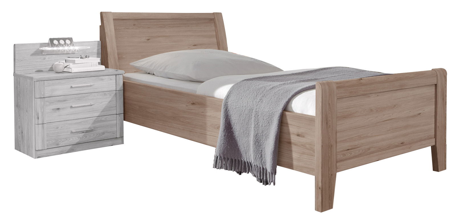 Komfortbett aus Holz mit grauer Decke und Kissen, seitliche Perspektive, daneben ein Nachttisch mit Schubladen.