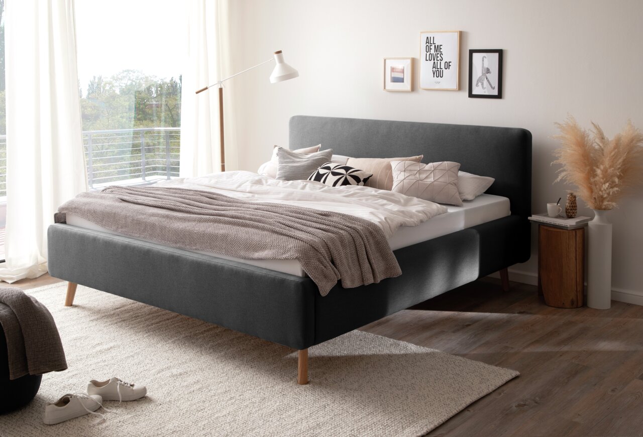 Modernes Polsterbett in einem hellen Schlafzimmer, seitliche Perspektive. Das Bett hat ein graues Kopfteil und Holzfüße, dekoriert mit Kissen und einer Decke.
