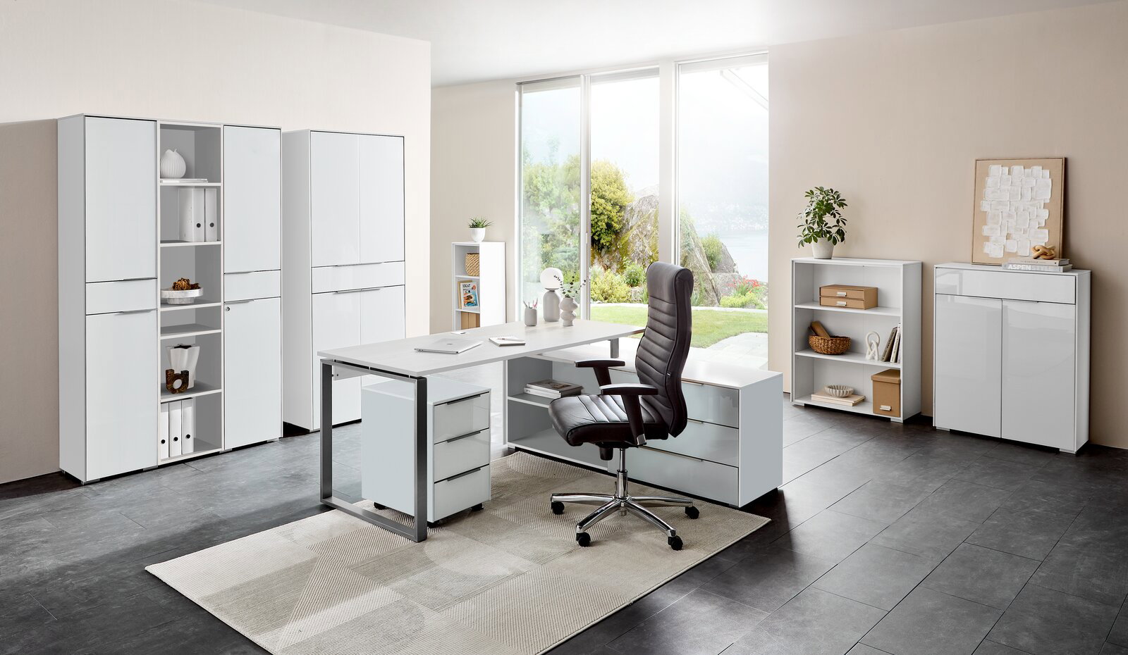 Voleo Aktenregal NEVADA Moderne Büromöbelkombination in einem hellen Raum mit großem Fenster. Im Vordergrund ein Eckschreibtisch mit Rollcontainer und ergonomischem Bürostuhl. Im Hintergrund mehrere Aktenschränke und Regale. Perspektive von vorne.
