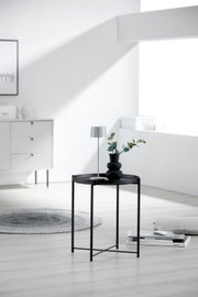 Schwarzer runder Beistelltisch aus Metall mit Tablettkante, darauf kleine Tischlampe und Vase mit Zweigen, im hellen Wohnzimmer – Frontansicht auf Augenhöhe