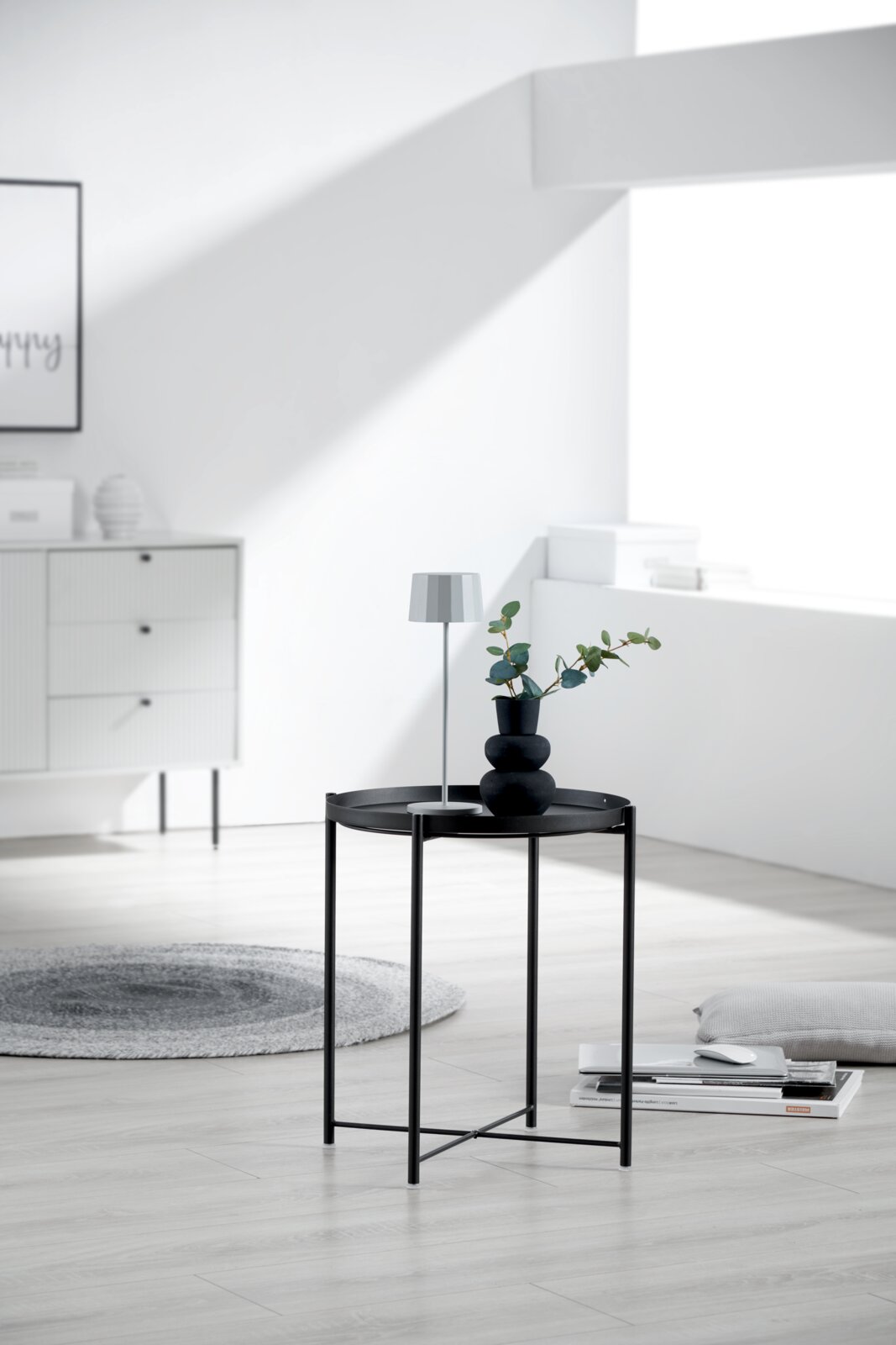 Schwarzer runder Beistelltisch aus Metall mit Tablettkante, darauf kleine Tischlampe und Vase mit Zweigen, im hellen Wohnzimmer – Frontansicht auf Augenhöhe
