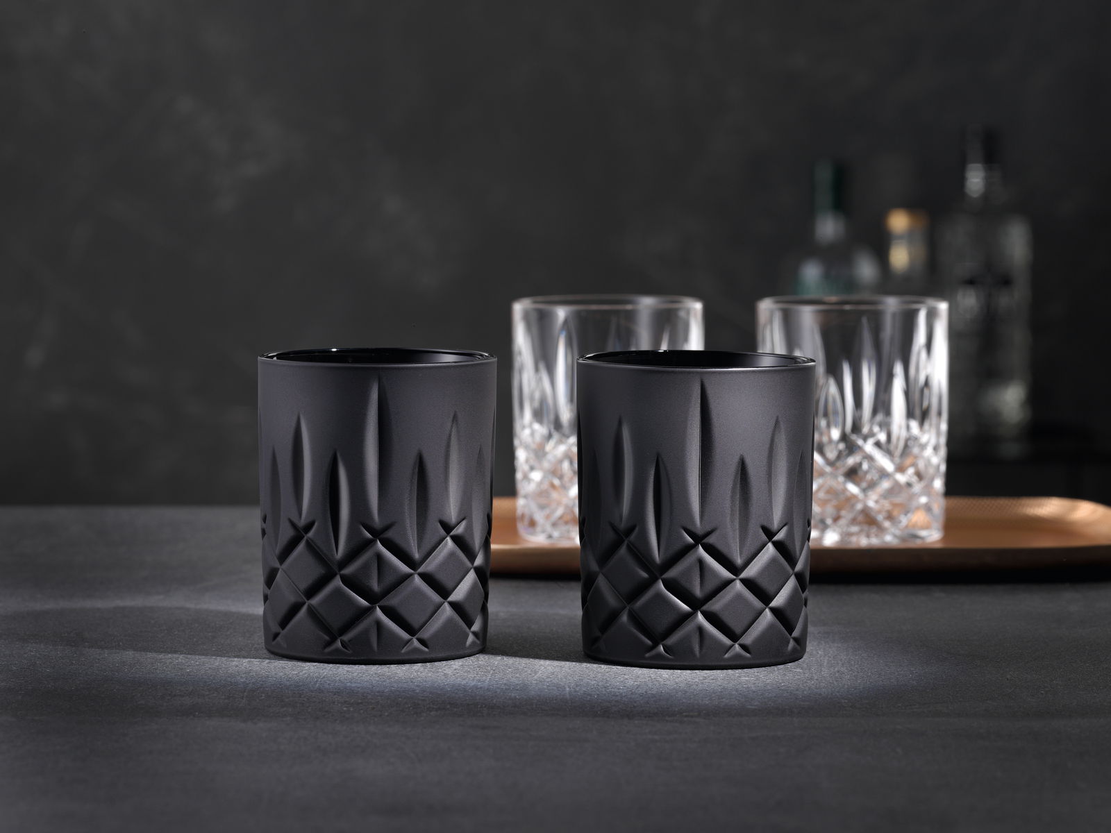 Zwei schwarze Whiskybecher mit elegantem, geometrischem Muster, frontal auf einem dunklen Hintergrund präsentiert.