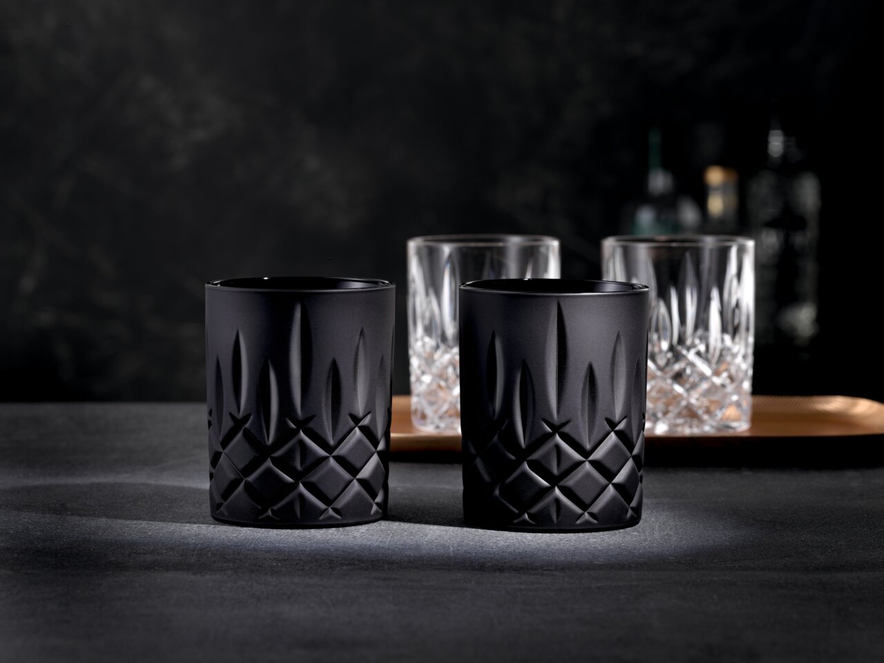Zwei schwarze Whiskybecher mit elegantem, geometrischem Muster, frontal auf einem dunklen Hintergrund präsentiert.