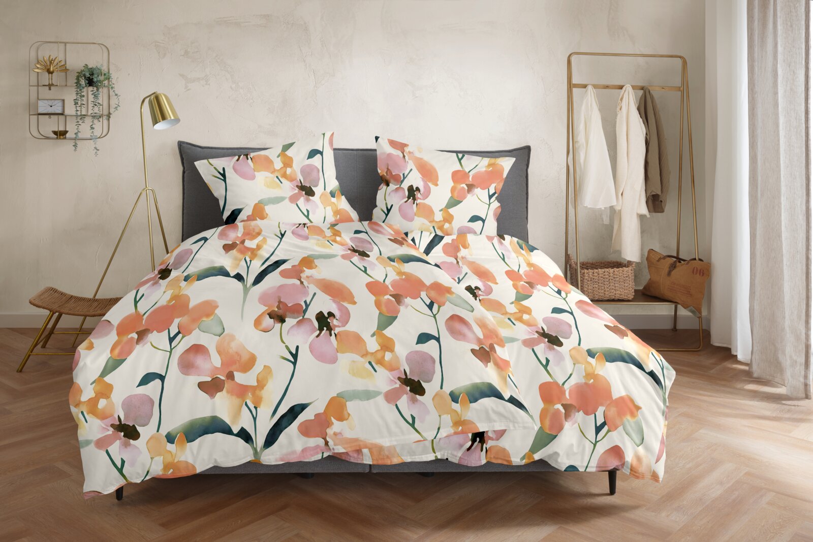 Blumige Bettwäsche mit Aquarell-Blütenmuster in Orange, Rosa und Grün auf weißem Grund, auf einem grauen Bett im Schlafzimmer – Frontansicht leicht von oben.
