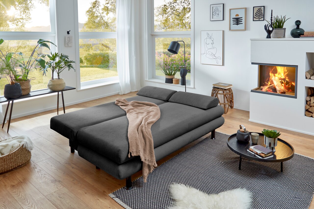 Modernes Funktionssofa in einem gemütlichen Wohnzimmer mit Blick auf den Garten, aus der Perspektive von vorne links.