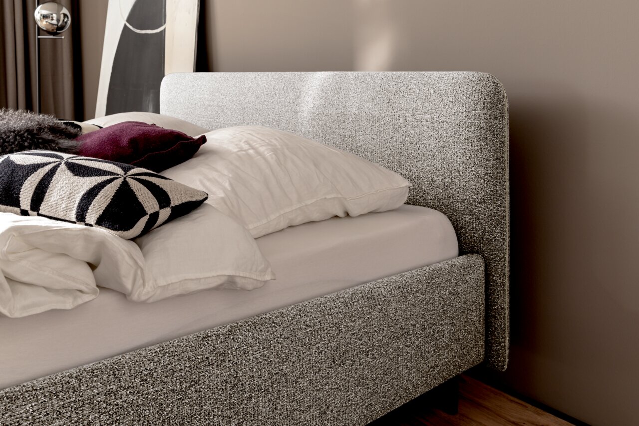 Zijaanzicht van een gestoffeerd bed met een grijze stoffen bekleding, versierd met witte en gedessineerde kussens.