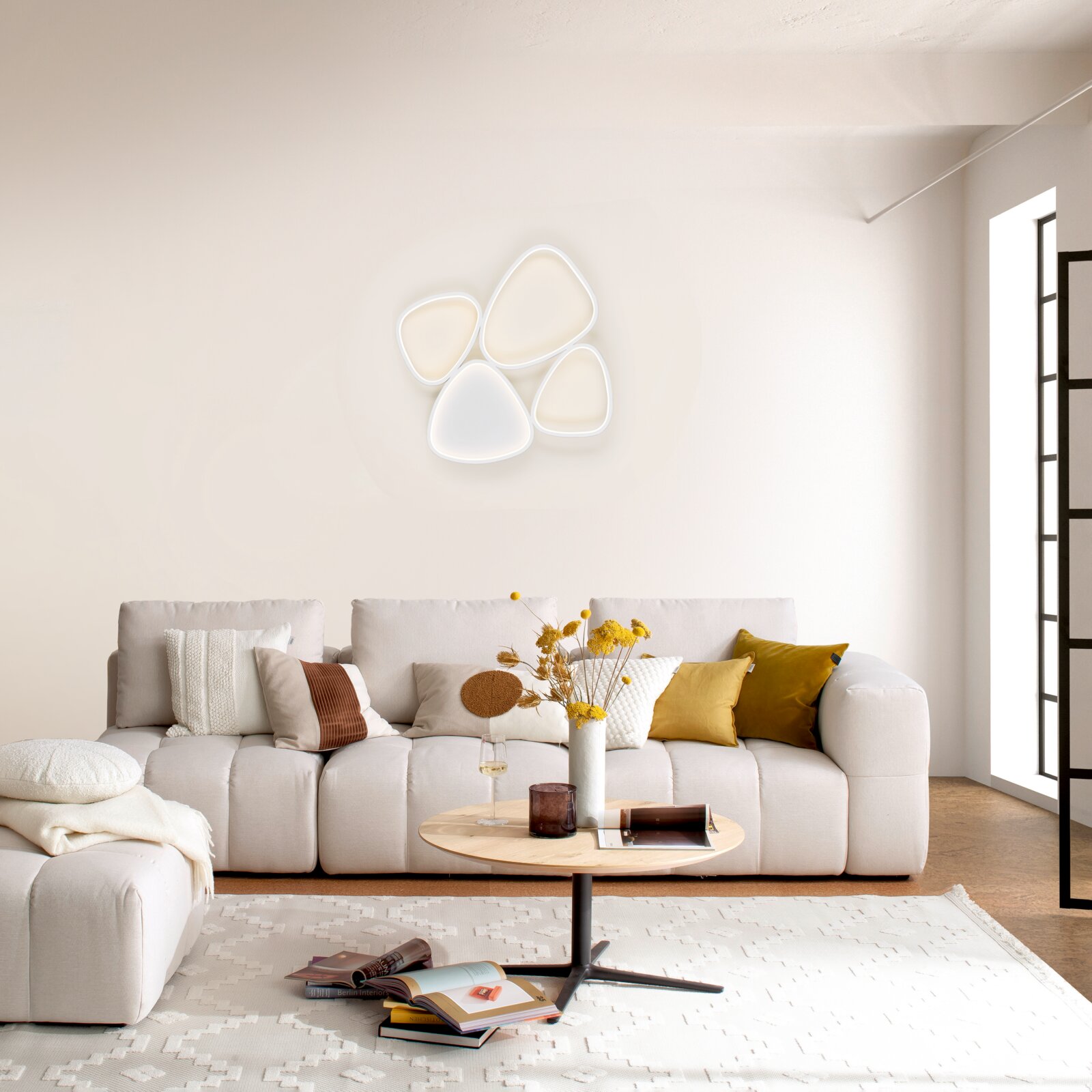 Moderne LED-Wandleuchte in organischer Kleeblatt-Form mit vier leuchtenden Elementen über einem hellen Sofa im Wohnzimmer, frontale Ansicht leicht von rechts
