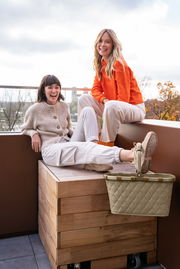Zwei lachende Frauen sitzen auf einer Holzbox auf einem Balkon, mit einem Carrybag in Rhombus Olive im Vordergrund.