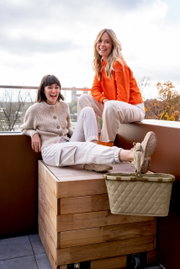Zwei lachende Frauen sitzen auf einer Holzbox auf einem Balkon, mit einem Carrybag in Rhombus Olive im Vordergrund.
