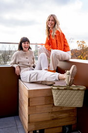 Zwei lachende Frauen sitzen auf einer Holzbox auf einem Balkon, mit einem Carrybag in Rhombus Olive im Vordergrund.