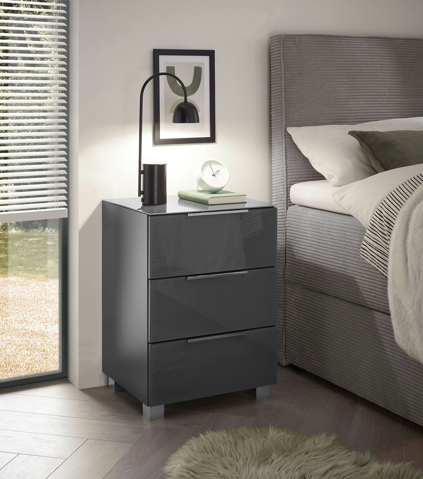Moderne Nachtkommode mit drei Schubladen in Hochglanzoptik, seitliche Perspektive neben einem Bett, dekoriert mit einer Lampe und Büchern.
