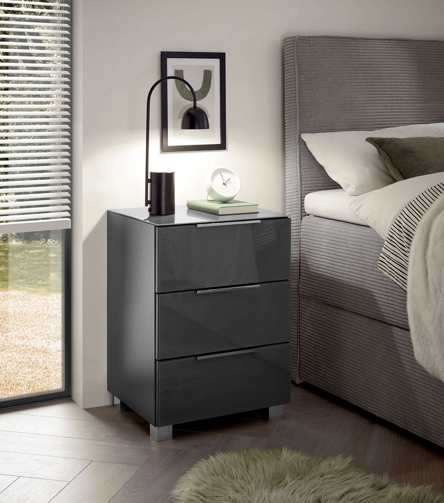 Moderne Nachtkommode mit drei Schubladen in Hochglanzoptik, seitliche Perspektive neben einem Bett, dekoriert mit einer Lampe und Büchern.