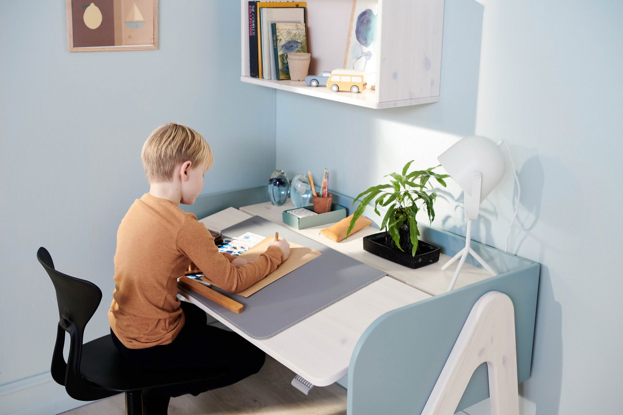 Ein Kind sitzt an einem Schreibtisch und malt auf einer Schreibtisch-Auflage. Der Schreibtisch ist hell und modern gestaltet, mit einer Lampe und einer Pflanze darauf. Die Perspektive zeigt die Szene von der Seite.