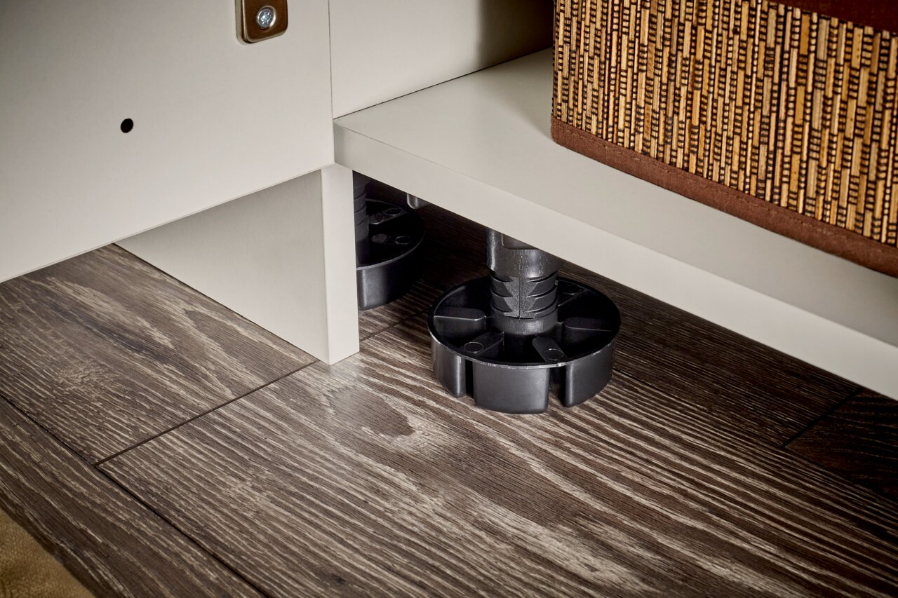 Nahaufnahme aus niedriger Perspektive: höhenverstellbarer schwarzer Möbelfuß unter einem weißen Schrank auf Holzfußboden