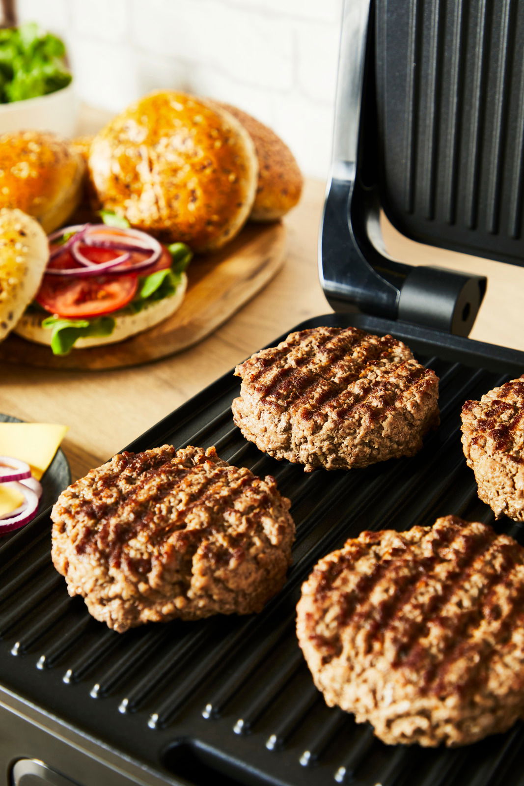 Kontaktgrill mit gegrillten Burger-Patties, im Hintergrund frische Burgerbrötchen und Zutaten, seitliche Perspektive